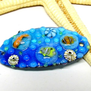 Könnte beinhalten: Ein ovaler, blauer Glasanhänger mit Meeresleben-Motiv. Der Anhänger zeigt bunte Fische, Muscheln und Blasen.