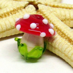 May include: A small glass mushroom charm with a red cap with white polka dots and a white stem with a green base.