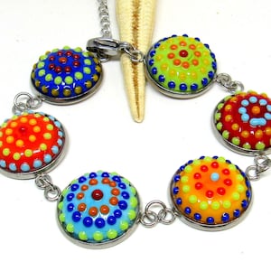 Könnte beinhalten: Ein farbenfrohes Armband mit sechs runden Glasperlen. Jede Perle hat ein anderes Muster aus kleinen Punkten in Rot-, Orange-, Gelb-, Grün-, Blau- und Brauntönen. Die Perlen sind durch eine silberne Kette verbunden.