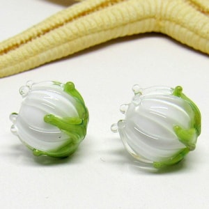 May include: Two white glass beads with green accents, resembling stylised flowers. The beads have a textured, ribbed surface and are perfect for jewellery making.