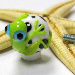 May include: A green and white glass frog knob with black polka dots. The frog has a blue eye and is holding its legs up. The knob is attached to a silver screw.