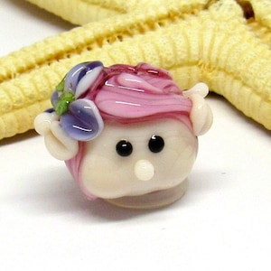 May include: A small glass bead with a pink and white swirl design, resembling a face with black eyes and a white nose. The bead is decorated with a purple and green flower.