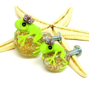 Könnte beinhalten: Zwei grüne Glas-Schubladengriffe in Froschform mit goldenem Glitzer und einer Krone aus schillernden Perlen. Die Froschgriffe sind an silbernen Schrauben befestigt.