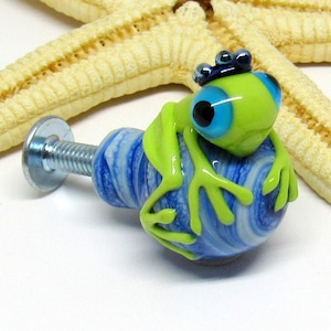 Könnte beinhalten: Ein grüner Glasfrosch mit blauen Augen und einer blauen Krone sitzt auf einem blauen und weißen, spiralförmigen Glasknopf. Der Knopf hat eine silberne Schraube.