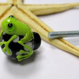 May include: A green glass frog figurine perched on a black and white polka dot glass sphere. The sphere has a screw on the bottom for attaching to a surface.