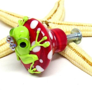Peut inclure: Un champignon en verre rouge et blanc avec des pois blancs et une grenouille verte assise dessus. La grenouille a un corps vert, des yeux blancs et une couronne argentée.