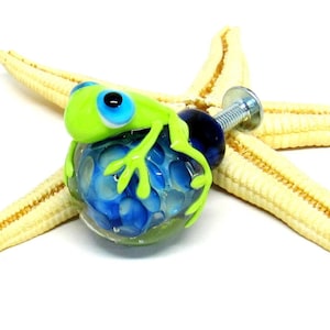 May include: A green glass frog figurine with blue eyes sits on top of a blue and white glass knob with a silver screw. The knob is on a starfish.