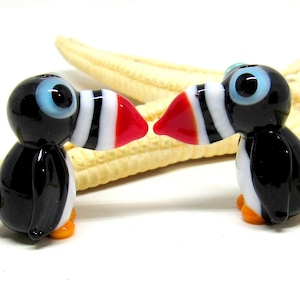 May include: Two small glass figurines of puffins, each with black and white striped beaks, red tips, and black bodies. The puffins are facing each other with their beaks touching, as if they are kissing. The figurines are sitting on a starfish.