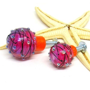 Könnte beinhalten: Zwei rosafarbene und orangefarbene Glas-Schrankknöpfe mit schwarzen Wirbeln und silbernen Akzenten. Die Knöpfe sind rund und haben eine strukturierte Oberfläche.