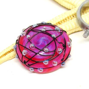 May include: A round, pink and purple glass bead with a black wire design and small, iridescent glass beads. The bead is approximately 1 inch in diameter. A starfish and metal hardware are in the background.