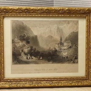 Pode incluir: Uma impressão de paisagem monocromática emoldurada que retrata um vale de montanha com uma pequena aldeia, edifícios de pedra e figuras com gado. A obra está inserida numa moldura dourada ornamentada e texturizada com uma margem creme.