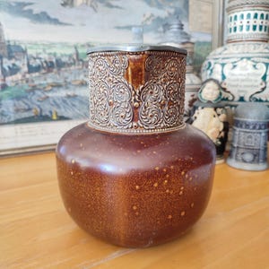 May include: A brown ceramic pitcher with a decorative silver-colored band around the top. The band features an ornate floral design. The pitcher has a rounded body and a small, flat lid. The background includes framed artwork and other decorative items.