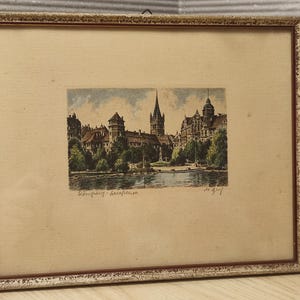 Pode incluir: Uma gravura emoldurada retratando uma paisagem urbana com uma torre de igreja alta, edifícios históricos e árvores ao longo de uma orla. A obra apresenta tons suaves de verde, castanho e azul. O texto "Königsberg - Schlosskirche" está escrito abaixo.