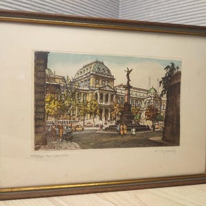 Puede incluir: Un grabado enmarcado que representa una plaza urbana con un gran edificio con cúpula y un monumento central con una estatua. La obra utiliza tonos apagados de verde, amarillo y marrón, e incluye texto escrito a mano en el margen inferior.