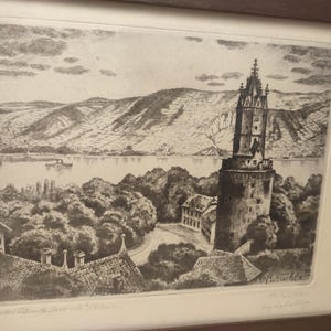 Pode incluir: Uma gravura a preto e branco numa moldura de madeira castanha representando uma paisagem com uma torre de pedra alta, árvores densas e um rio que flui junto a colinas. O texto "Blick der Turm/Bacharach a/Rhein" está inscrito no canto inferior esquerdo.