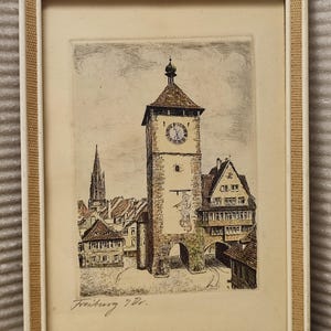 Puede incluir: Un grabado enmarcado de la puerta Schwabentor en Friburgo, Alemania, que muestra una torre de reloj central con una figura pintada, edificios circundantes y la aguja de una iglesia a lo lejos. Firmado Freiburg i. Br. en la esquina inferior izquierda.