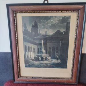 May include: Framed print of an architectural scene with a fountain, arches, and figures. The print is in shades of gray and brown, set within a dark brown wooden frame with a decorative gold trim. A metal ring is attached for hanging.