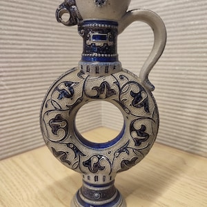 Può includere: Una brocca in ceramica circolare con un design centrale ad anello aperto, decorata con motivi floreali blu scuro e grigi. Il recipiente presenta un manico curvo, un bordo svasato con un piccolo beccuccio e una base a piedistallo con dettagli in rilievo.