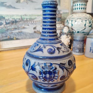 Pode incluir: Uma garrafa de cerâmica azul e cinza com um design detalhado. A garrafa apresenta um pescoço texturizado e canelado e um corpo bulboso adornado com motivos florais e de brasão. É provavelmente uma peça decorativa ou um recipiente.