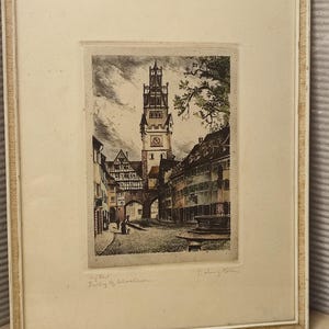 May include: A framed etching depicting a town square with a tall clock tower, half-timbered buildings, and a fountain. The artwork features muted tones and includes handwritten text at the bottom left and a signature at the bottom right.