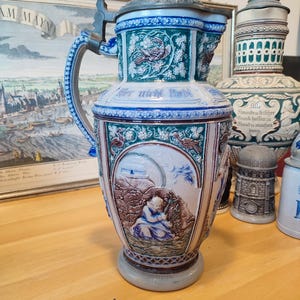 Puede incluir: Una jarra de cerveza de cerámica grande y ornamentada con tapa metálica abisagrada. La jarra presenta diseños en relieve detallados en tonos azules, verdes y marrones, incluyendo patrones florales y una escena de una persona. El asa es azul y blanca.