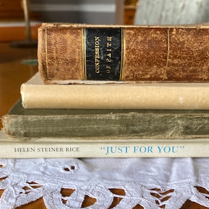 May include: A stack of vintage books with aged covers. The top book, "CONFESSION OF FAITH," has a dark spine. Below are books in cream and olive green. The bottom book, "JUST FOR YOU," rests on a white lace doily.