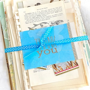 May include: A collection of vintage ephemera, including old book pages, maps, and a blue card with "THANK YOU" in gold lettering, tied with a blue polka dot ribbon. The pages feature illustrations of fish and other marine life.