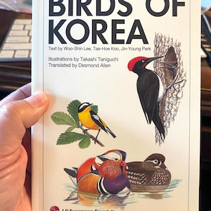 May include: A field guide titled "Birds of Korea" featuring illustrations of various bird species. The cover displays a yellow and black bird on a branch, a black and white woodpecker, and colorful mandarin ducks. Text includes author and translator names.