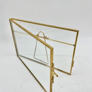 May include: A gold-coloured metal and glass photo frame, open and propped up with a small metal stand. The frame is rectangular with a hinged design, allowing it to stand upright. The glass panels are clear.