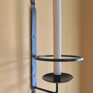 May include: A black metal wall-mounted candle holder with a white candle. The holder has a circular ring and a small, round base to catch wax. The design includes decorative leaf-shaped accents.