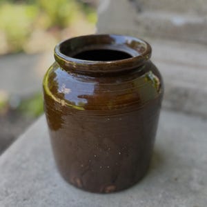 Pode incluir: Um pote de cerâmica vidrada em castanho escuro com uma boca larga e um rebordo espesso e arredondado. O recipiente cilíndrico apresenta um acabamento liso e brilhante com variações subtis de cor e uma base ligeiramente cónica.