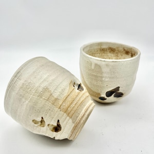 May include: Two handmade ceramic cups with a textured, off-white glaze and brown accents. One cup is tilted on its side, revealing the interior. The other cup is upright, showcasing the rim and interior. The cups have a rustic, artisanal aesthetic.