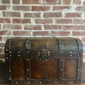 Op de afbeelding: Een donkerbruine houten kist met metalen details. De kist heeft een afgeronde bovenkant en is vastgezet met metalen banden en klinknagels. Een decoratieve metalen sluiting is gecentreerd aan de voorkant. De kist staat op een donker houten oppervlak.