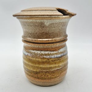 May include: A handcrafted ceramic jar with a lid. The jar features a cylindrical body with horizontal ridges and a flared top. The color palette includes shades of brown, tan, and gold, with a subtle white and blue stripe. The lid has a small opening.