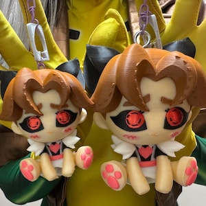 以下が含まれることがあります： 2つのアニメスタイルのキャラクターキーホルダー。各キーホルダーは、茶色の髪、黒いリボン、赤い目を持つ人形が特徴です。人形はピンクのアクセントが付いた白と黒の衣装を着ており、足にはピンクの足跡があります。銀色のチェーンから吊り下げられています。