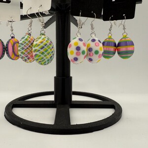 May include: A set of six pairs of colorful Easter egg earrings. The earrings are made of polymer clay and feature various patterns, including polka dots, stripes, and geometric designs. The earrings are displayed on a black earring stand.