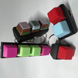May include: A set of four keychains featuring different colored keycaps. Each keychain has a black plastic frame and a metal ring. The keycaps are made of plastic and have a textured surface. The keycaps are in the colors pink, green, blue, and red.