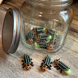 May include: A glass jar with a metal lid filled with small orange and black firefly figurines. Three more fireflies are sitting on a wooden surface outside the jar.