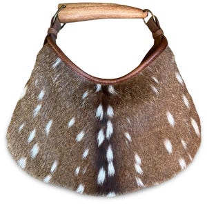 May include: A crescent-shaped handbag crafted from brown and white spotted animal hide. The bag features a smooth, curved wooden handle attached with brown leather loops and a dark brown leather trim along the top opening edge.