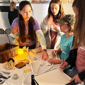 Puede incluir: Cuatro jóvenes están reunidos alrededor de una mesa, trabajando en un proyecto. Están usando un mapa, un cuaderno y una vela. Hay una cesta, pan y una taza pequeña en la mesa. La mesa está cubierta con un mantel blanco.