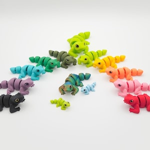 May include: A set of 11 colorful articulated toy lizards, each with a different color, arranged in a circle on a white background. The lizards are made of plastic and have movable limbs.