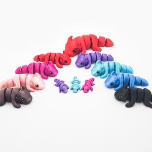 May include: A set of nine small, segmented, colorful, dragon-like figurines. The figurines are arranged in a circle, with the largest in the center and the smallest on the outside. The colors of the figurines are red, pink, purple, blue, and black.