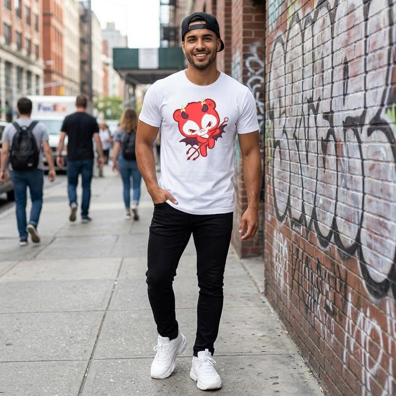 K&ouml;nnte beinhalten: Wei&szlig;es T-Shirt mit Rundhalsausschnitt und roter Teufel-Cartoon-Grafik. Die Person tr&auml;gt eine schwarze Jeans, wei&szlig;e Turnschuhe und eine schwarze Baseballkappe. Der Hintergrund zeigt eine Stadtstra&szlig;e mit Fu&szlig;g&auml;ngern und eine Backsteinmauer.