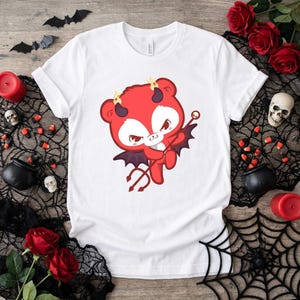 May include: White t-shirt featuring a cartoon devil bear with red fur, horns, and a trident. The bear has bat wings and a stern expression. The design is centered on the shirt, perfect for Halloween or themed events.