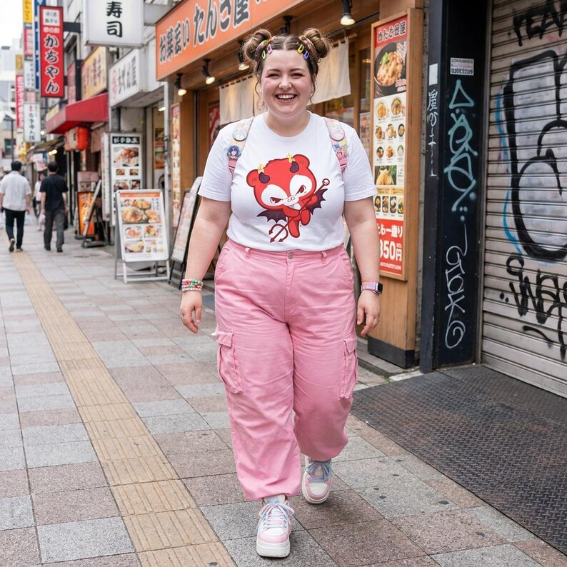 K&ouml;nnte beinhalten: Eine Person in einem wei&szlig;en T-Shirt mit einem roten Cartoon-Teufel-Motiv, rosa Cargohosen und rosa Turnschuhen geht eine Stadtstra&szlig;e entlang. Die Person hat ihre Haare in Dutt mit bunten Clips. Der Hintergrund zeigt Gesch&auml;fte und Schilder.