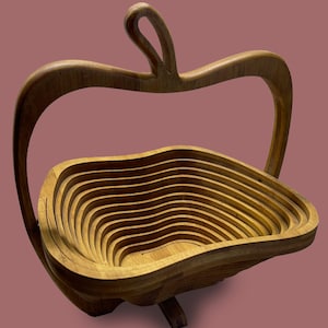 Puede incluir: Cesta de frutas de madera con forma de manzana, con asa. La cesta está hecha de madera en capas, creando un diseño interior ondulado. El color natural de la madera le da un aspecto rústico y elegante. La cesta está diseñada para colocarse sobre una superficie plana.