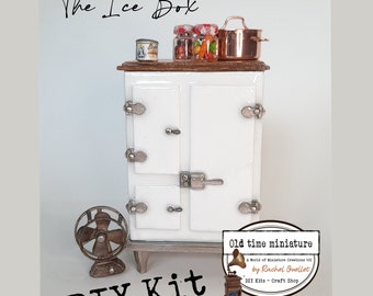 May include: A white miniature ice box with a wooden top and silver hardware. The ice box is sitting on a wooden base. There are jars of candy, a can of food, and a copper pot on top of the ice box. A small metal fan is in the foreground. The text "The Ice Box" is at the top of the image. The text "DIY Kit" is at the bottom of the image. The text "Old time miniature" is in a circle at the bottom right of the image. The text "A World of Miniature Creations 112 by Rachel Ouellet, DIY Kits - Craft Shop" is inside the circle.