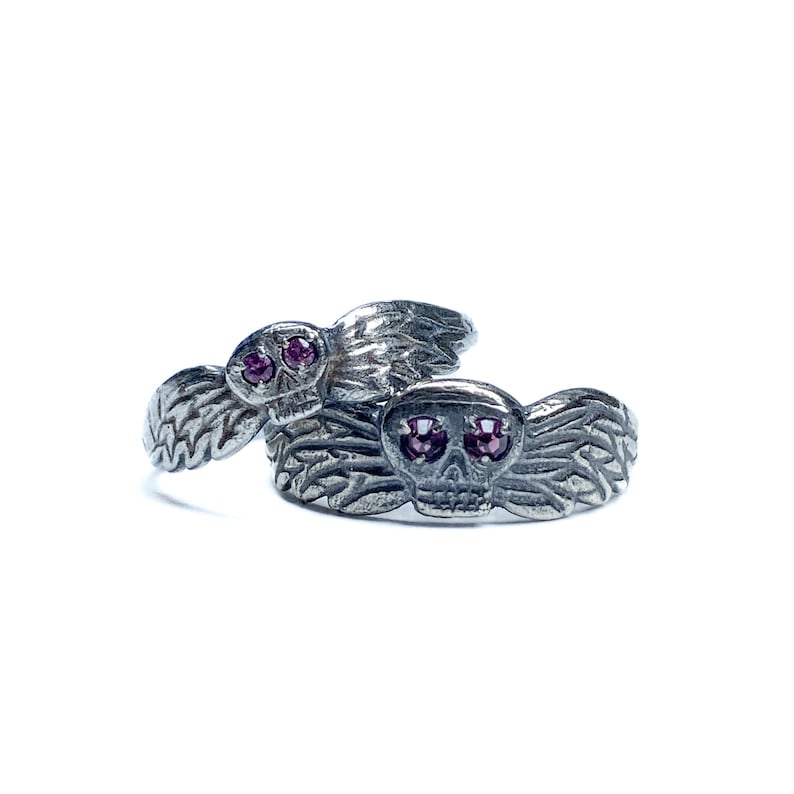 Puede incluir: Dos anillos de plata con forma de calavera con ojos de gemas rosas y alas. Los anillos est&aacute;n apilados uno encima del otro.