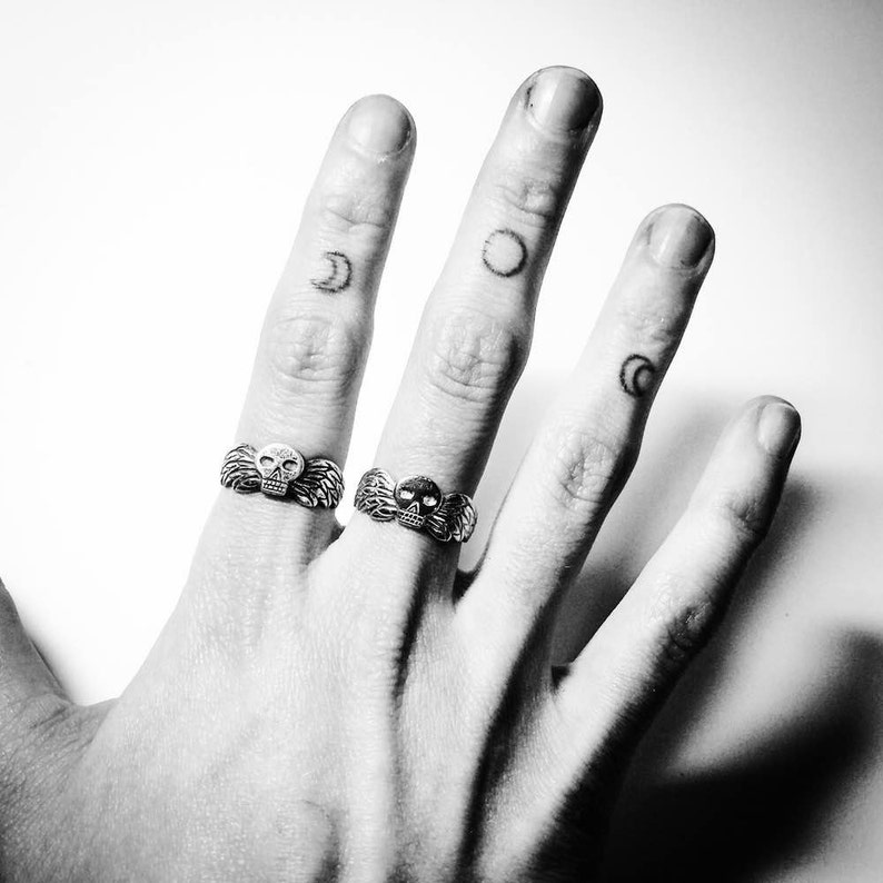 May include: A hand with two silver rings featuring skull designs. The rings are worn on the index and middle fingers. The hand also has several small crescent moon tattoos on the fingers.