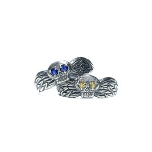 Puede incluir: Dos anillos de plata con forma de calavera y alas. Una calavera tiene ojos azules y la otra tiene ojos amarillos.